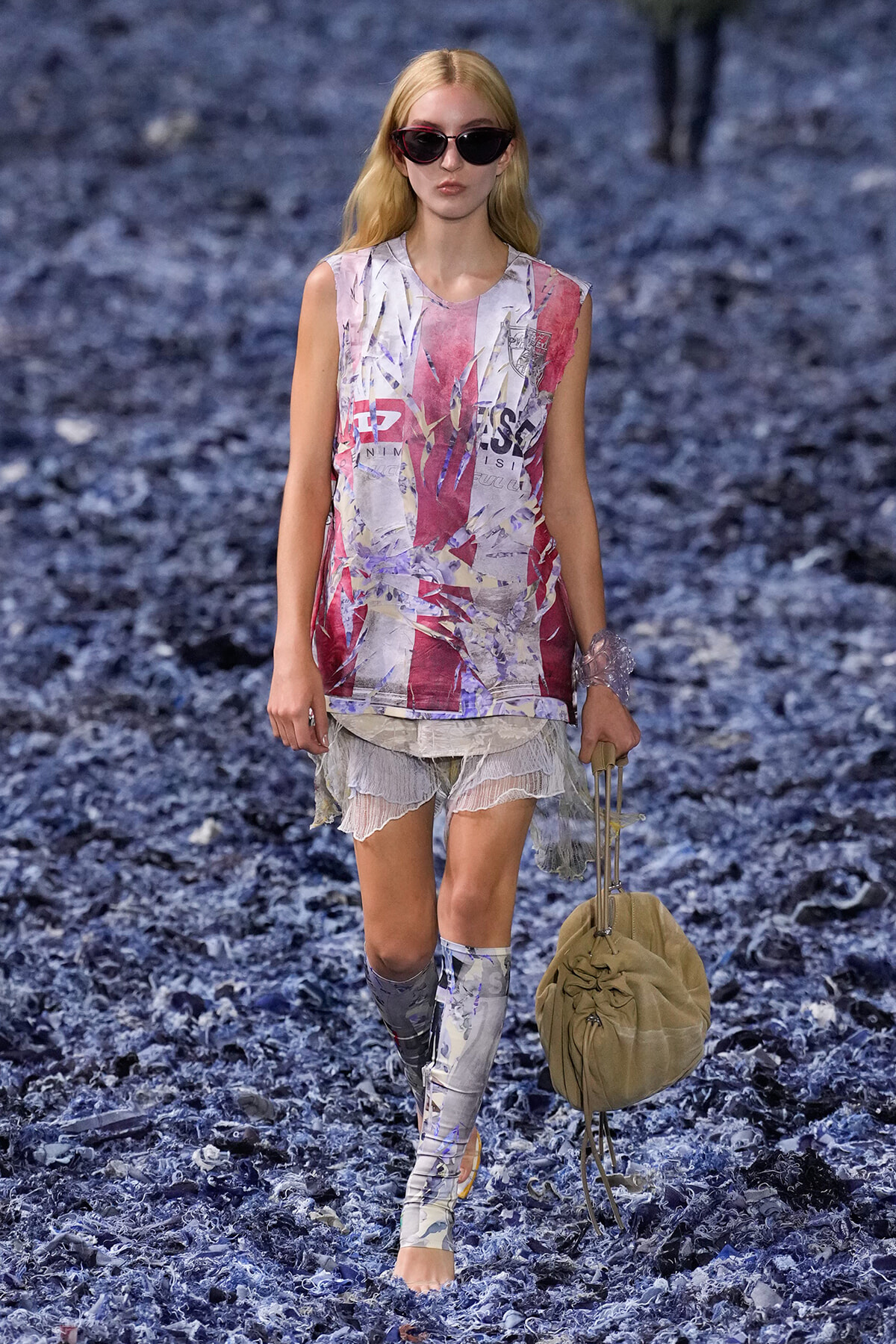 Model walking a runway on a blue-textured floor, wearing a sleeveless abstract pink–purple graphic top, sheer layered lace shorts, printed leg sleeves, red sunglasses, and a beige drawstring bag.