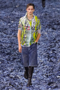 Male fashion model walks a runway wearing a lime-green, graphic-patterned shirt, blue denim knee-length shorts, and black knee-high boots.