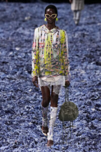Black model walking a fashion runway in a vibrant abstract print top, sheer lace shorts, and tall patterned boots, accessorized with neon yellow earrings and oversized sunglasses, carrying an olive green bag on the side.