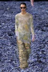 Model walking a runway in a sheer pastel floral top and heavily fringed, shredded pants with dark sunglasses.