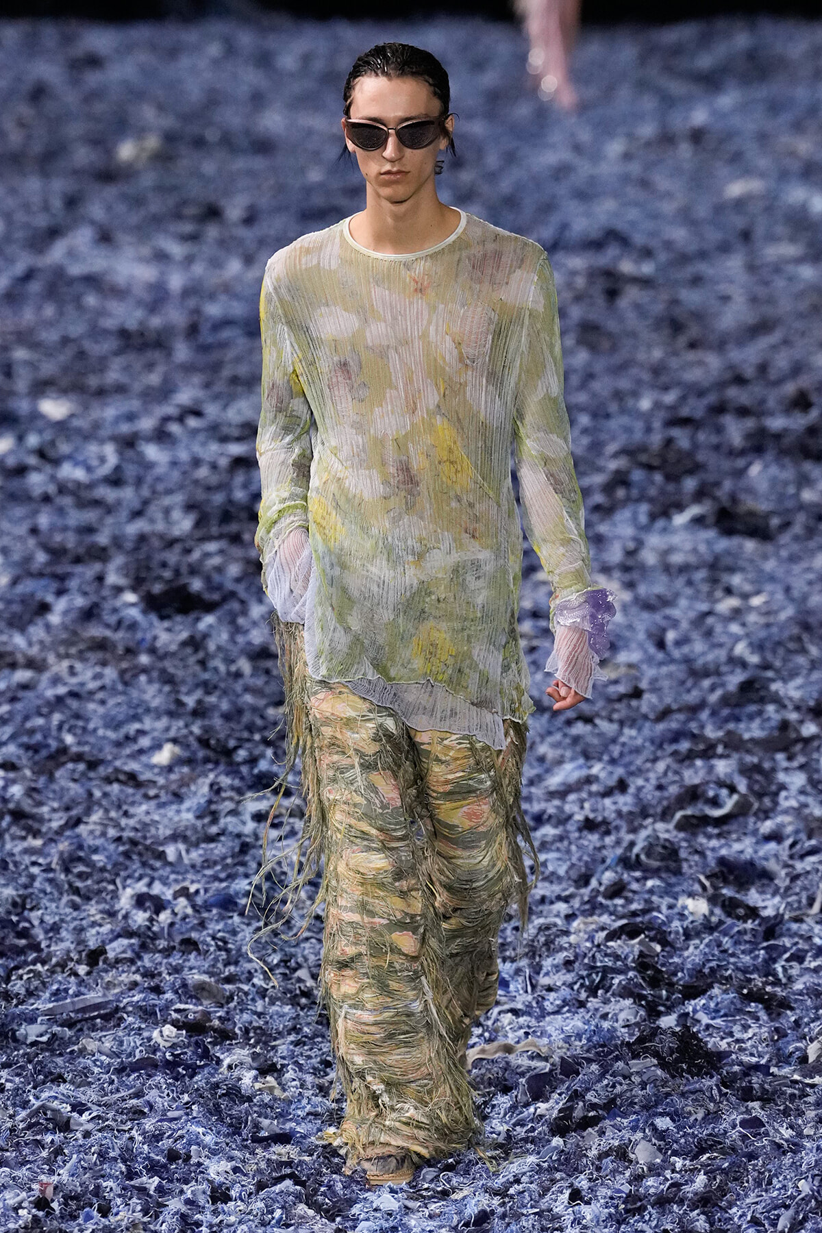 Model walking a runway in a sheer pastel floral top and heavily fringed, shredded pants with dark sunglasses.