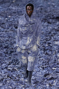 Model in an oversized, distressed blue-gray outfit with shredded fabric, walking on a runway covered in blue debris.