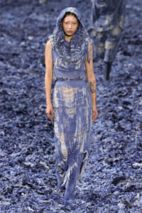 Model in a heavily distressed blue denim outfit with a hood, walking a runway covered in shredded blue material.