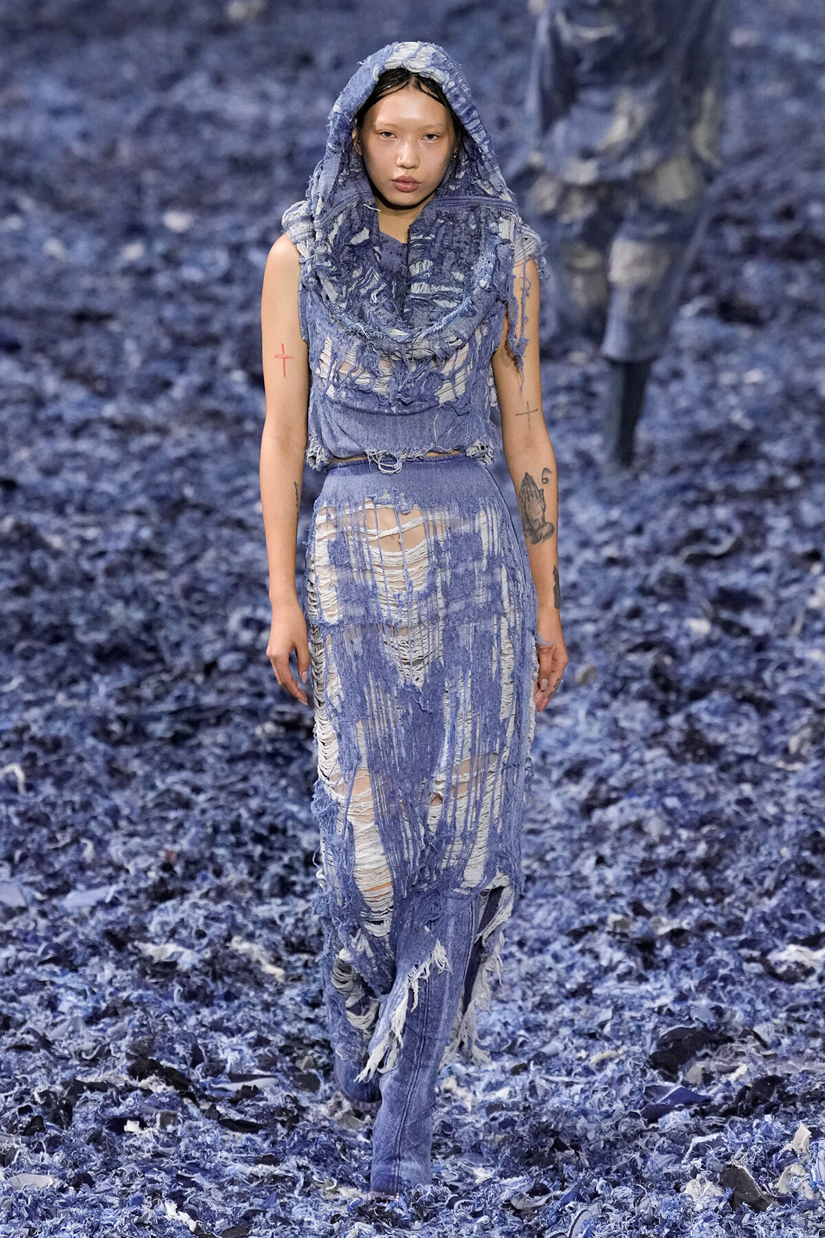 Model in a heavily distressed blue denim outfit with a hood, walking a runway covered in shredded blue material.