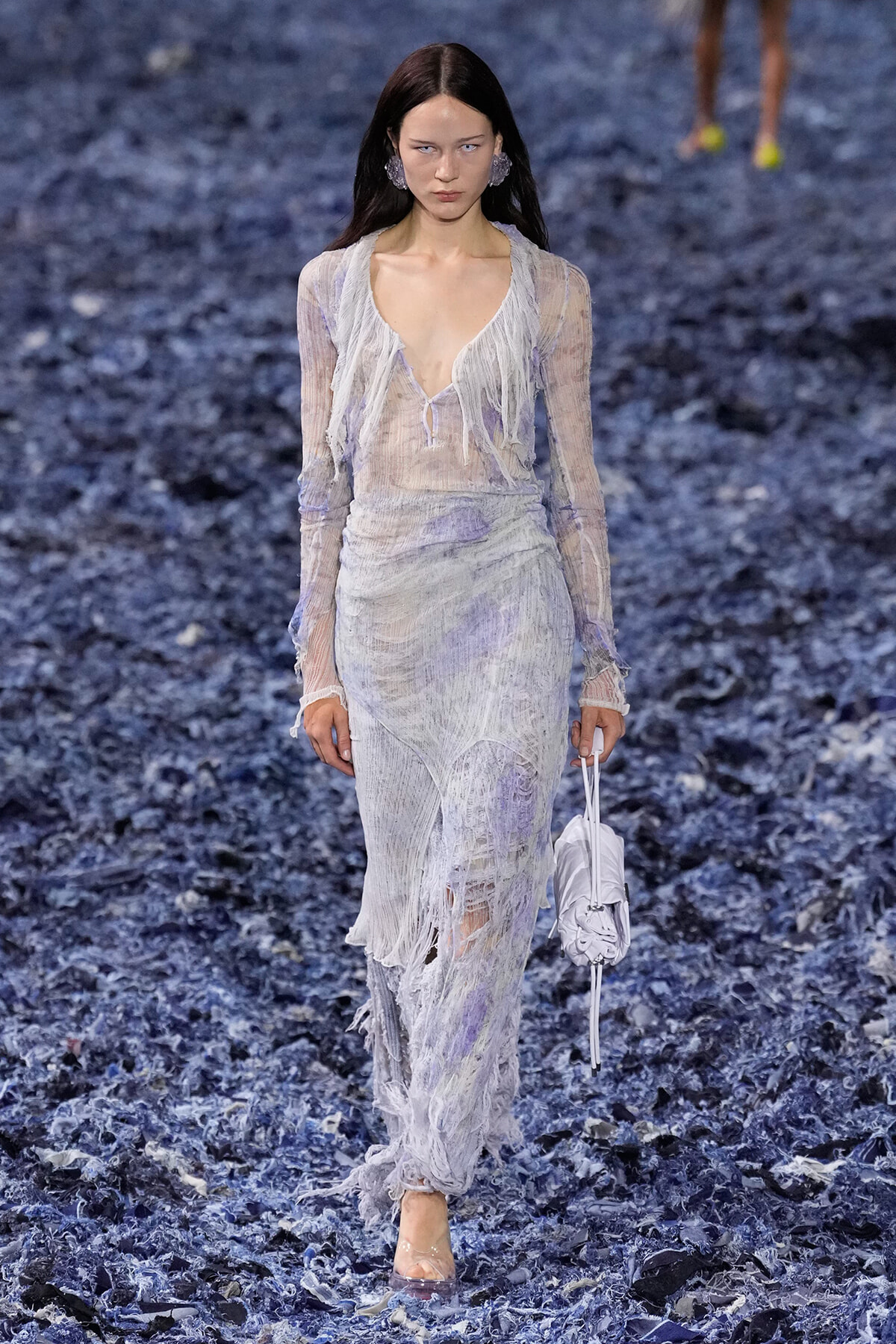Model walks a fashion runway wearing a sheer lavender-gray fringe dress with a deep neckline and long sleeves, carrying a small white bag.