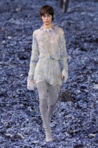 Female model walks the runway in a sheer, pleated blouse with a ruffled collar and light grey pants, carrying a gray handbag.