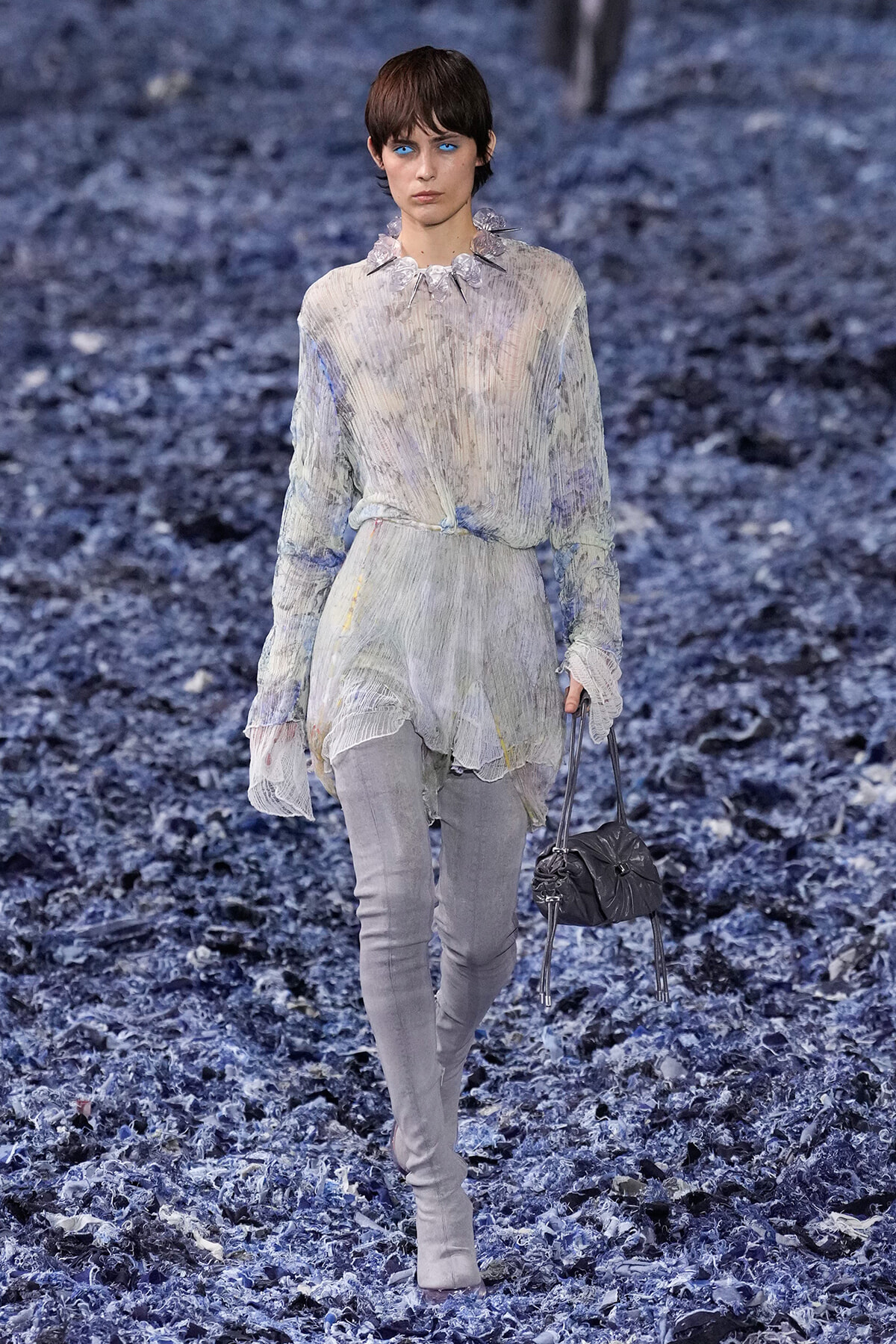 Female model walks the runway in a sheer, pleated blouse with a ruffled collar and light grey pants, carrying a gray handbag.