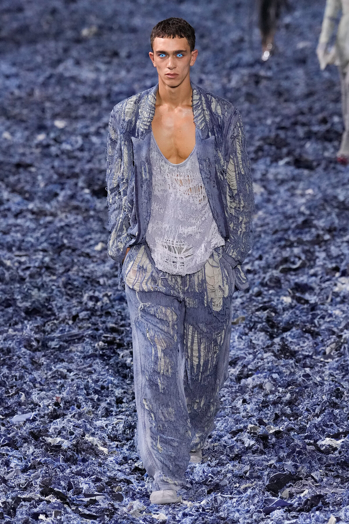 Male model in a distressed blue denim suit with a torn tank top walking a runway covered in shredded blue fabric groundfalling textures.