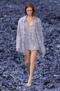Model walks the runway in a pale lavender lace set: oversized cardigan over a sheer, fringed dress with clear sandals.