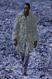 Male model walks toward the camera wearing a shaggy, pale blue-gray textured coat over blue jeans, in a rocky blue-toned landscape.