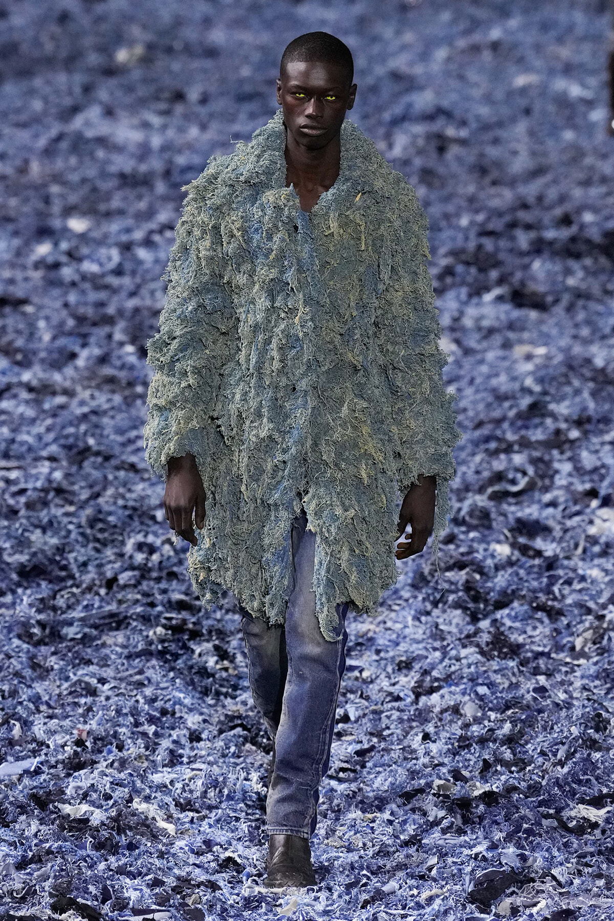 Male model walks toward the camera wearing a shaggy, pale blue-gray textured coat over blue jeans, in a rocky blue-toned landscape.