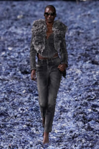 Model walking on a blue-gray rocky path, wearing a distressed denim outfit with a voluminous textured vest and dark sunglasses
