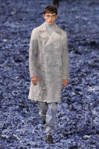 Male model walks a runway in a light gray, textured knee-length coat over a turtleneck, distressed jeans, and brown boots on a blue-toned runway surface.