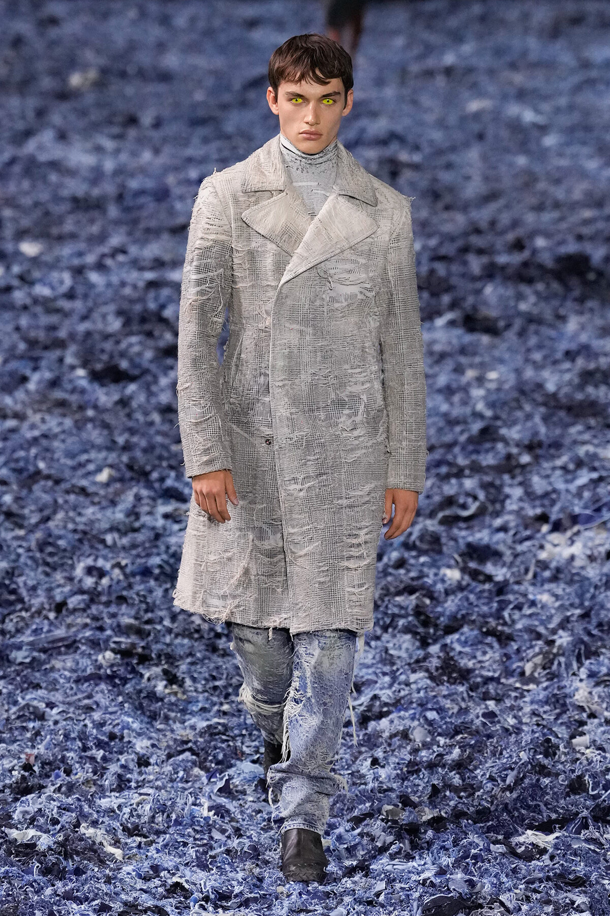 Male model walks a runway in a light gray, textured knee-length coat over a turtleneck, distressed jeans, and brown boots on a blue-toned runway surface.