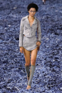 Model walking the runway in a distressed light gray two-piece outfit with a cropped jacket and mini skirt, paired with gray leg warmers and glasses on a blue-textured catwalk.