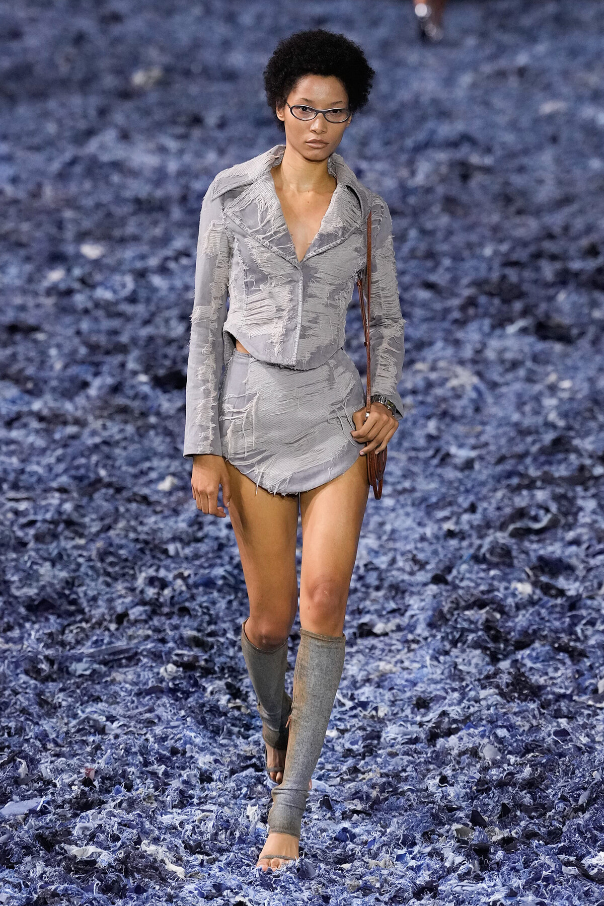 Model walking the runway in a distressed light gray two-piece outfit with a cropped jacket and mini skirt, paired with gray leg warmers and glasses on a blue-textured catwalk.