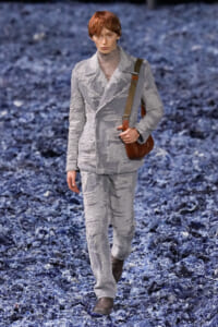 Male model walking a fashion runway in a distressed light-gray suit with frayed edges and a brown crossbody bag.