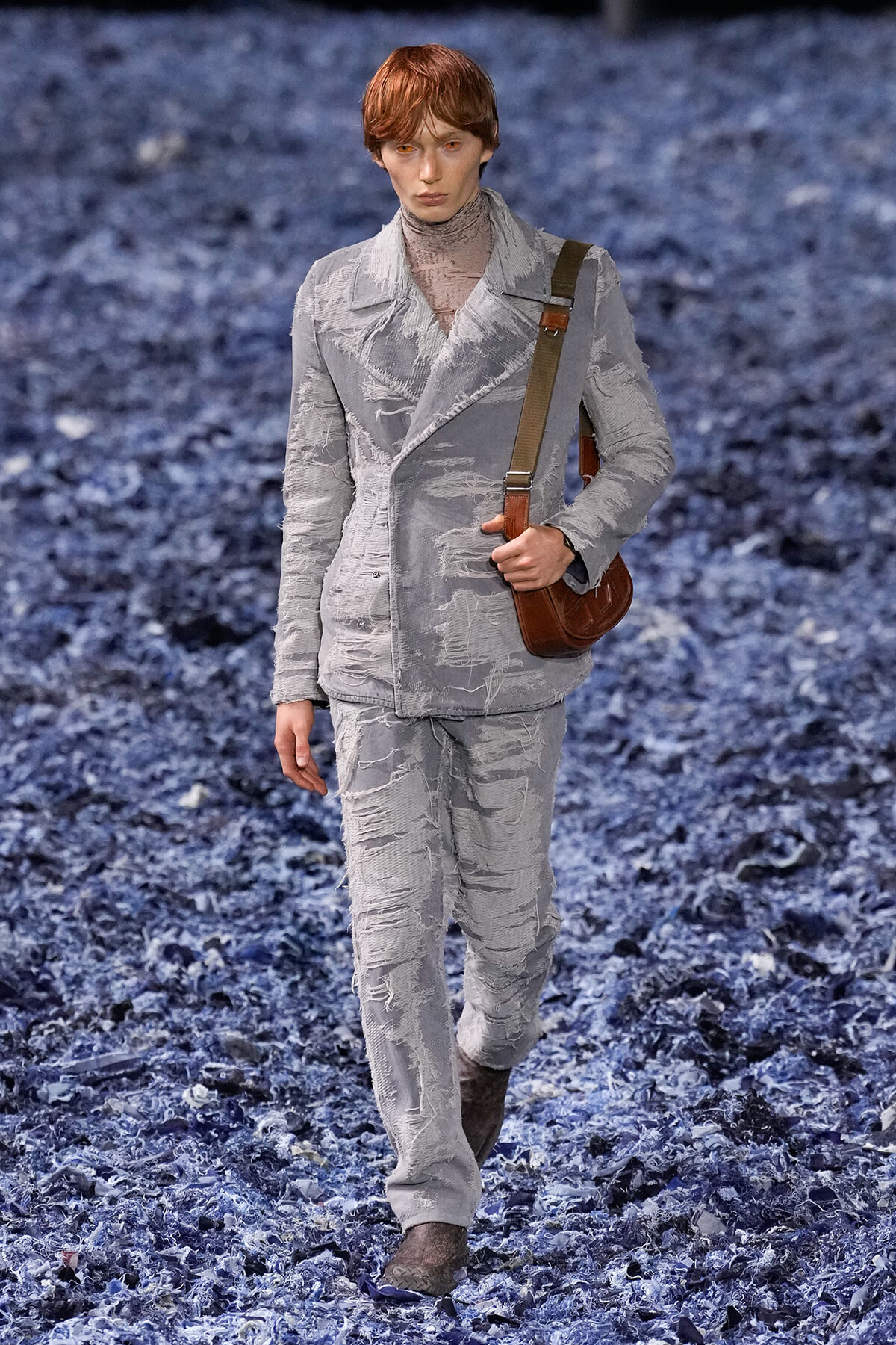 Male model walking a fashion runway in a distressed light-gray suit with frayed edges and a brown crossbody bag.