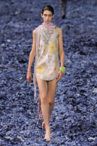Model walking a runway in a sleeveless floral mini dress with a pink striped scarf and fringe detail, on a blue-toned, textured runway surface.