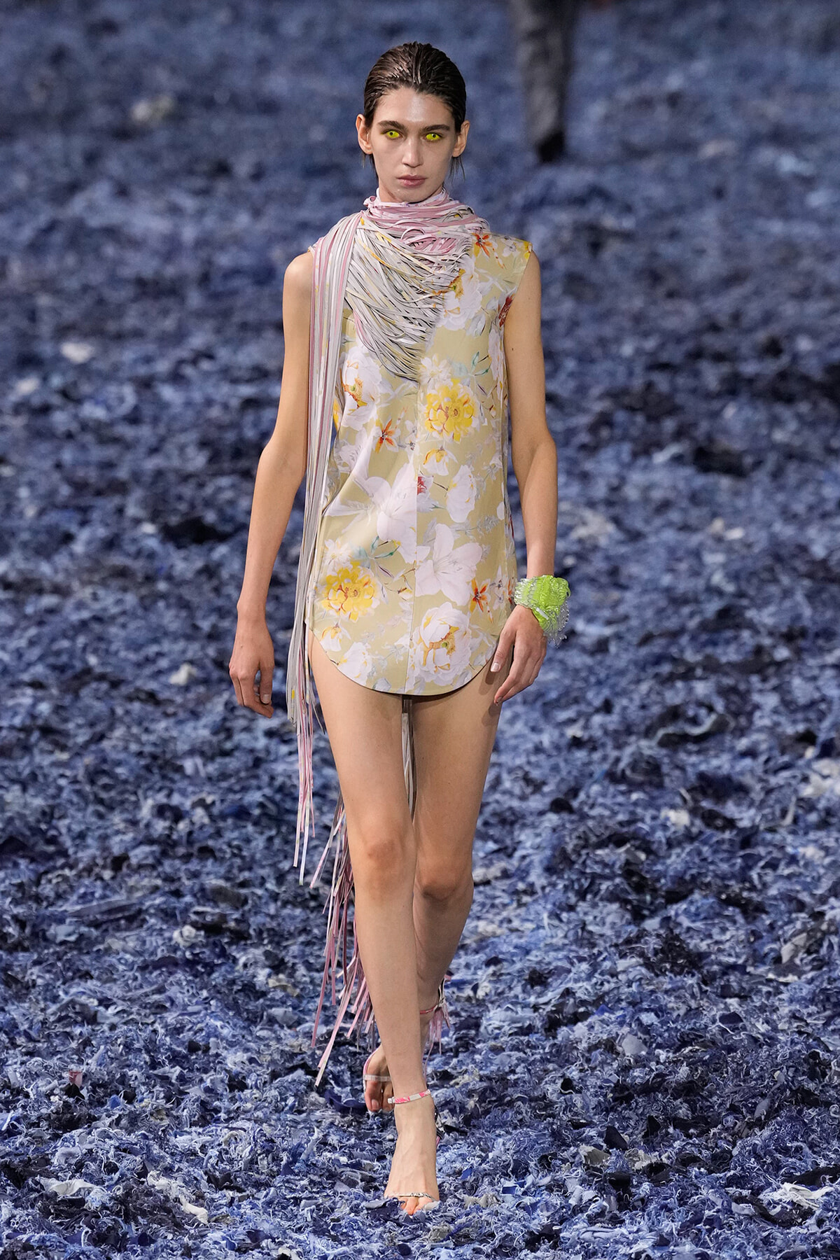 Model walking a runway in a sleeveless floral mini dress with a pink striped scarf and fringe detail, on a blue-toned, textured runway surface.