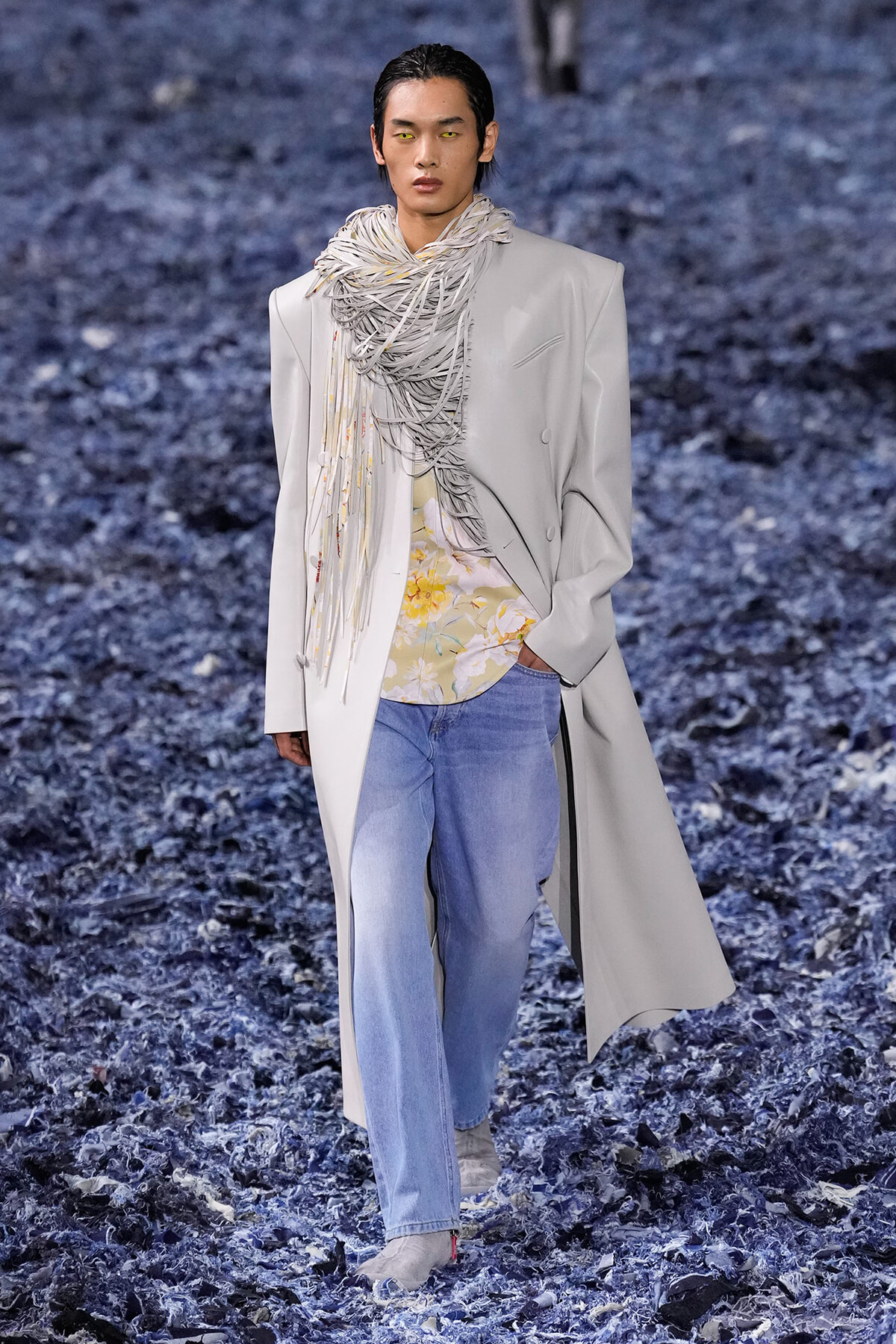 Male model walks a runway in a light gray overcoat with a sculptural scarf, floral shirt, and light-wash jeans on a blue-toned runway floor.