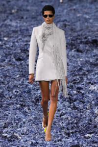 Female model walks a light gray blazer dress with a long fringe scarf and sunglasses on a blue textured runway.