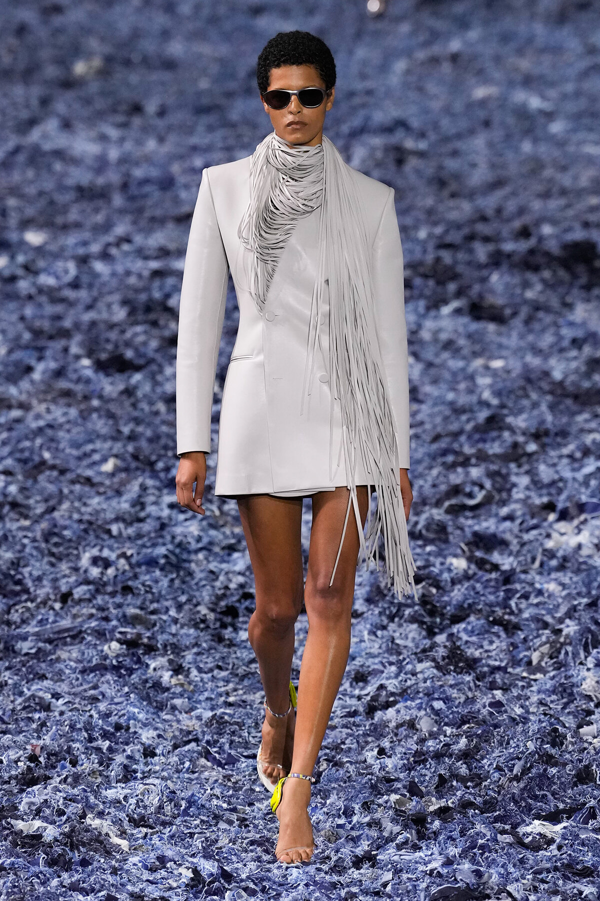 Female model walks a light gray blazer dress with a long fringe scarf and sunglasses on a blue textured runway.
