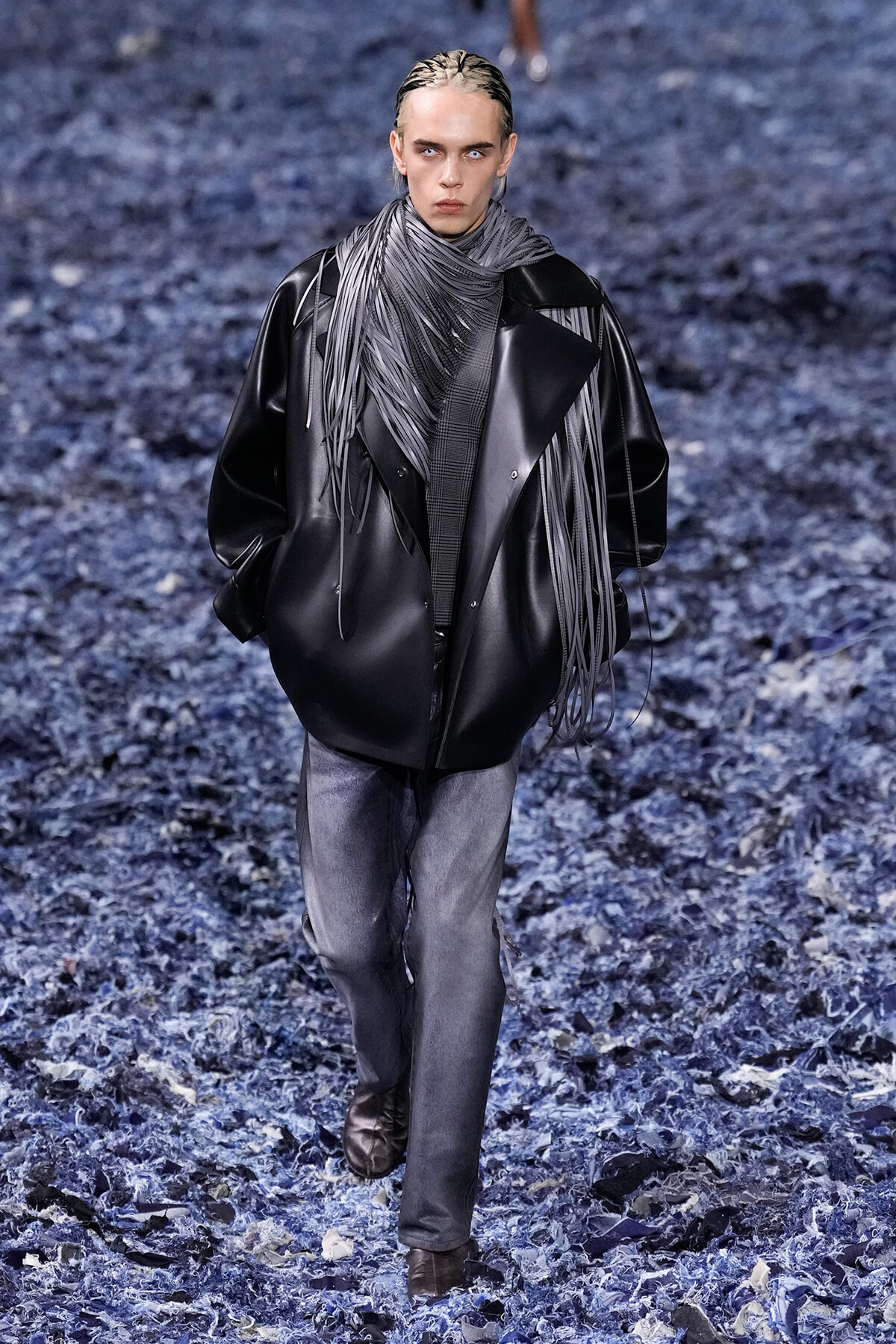 Model walks a runway in an oversized black leather coat with a long gray fringe scarf, gray jeans, and brown boots.