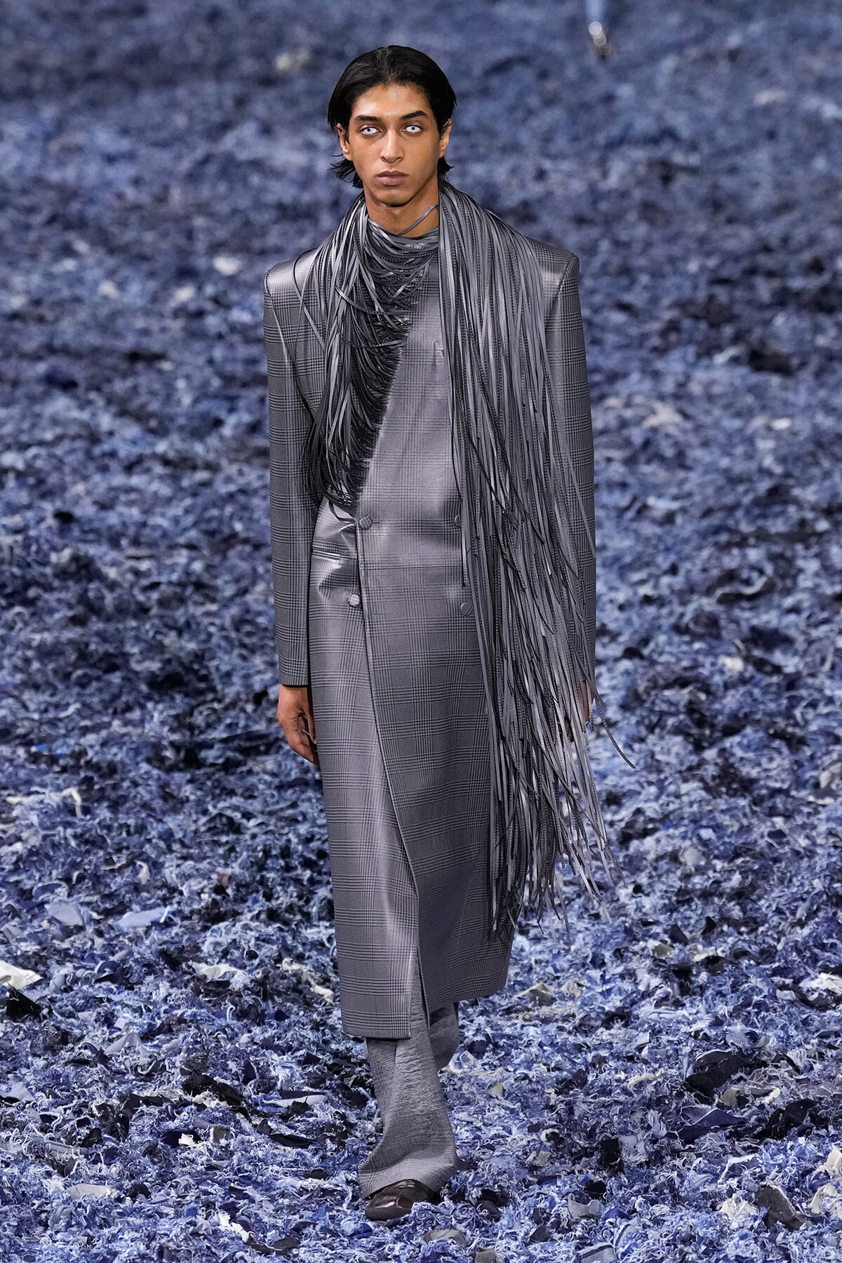 Male model walking a gray plaid double-breasted suit with a long, black fringe scarf on a blue-toned textured runway.