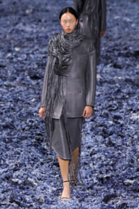 Model walks the runway in a gray plaid suit with a dramatic, wrapped fringe scarf cascading around the neck.