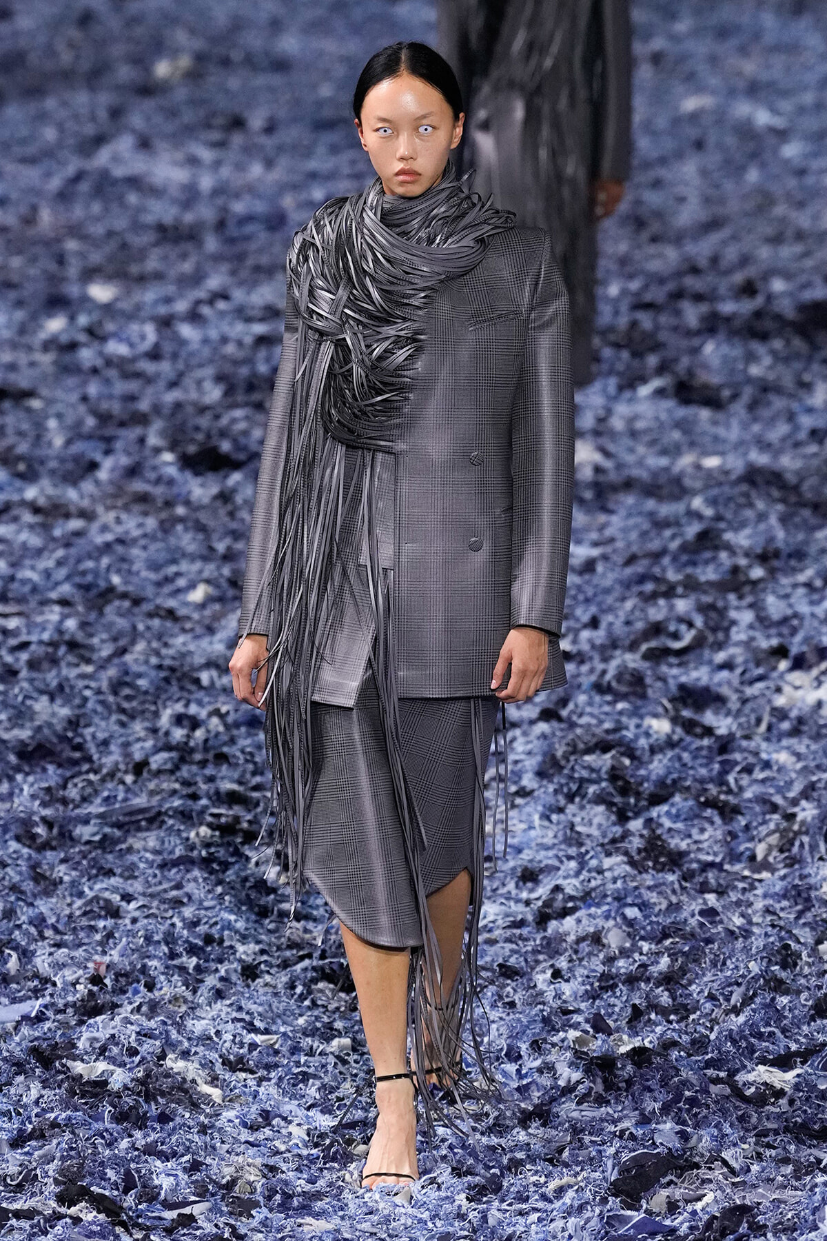 Model walks the runway in a gray plaid suit with a dramatic, wrapped fringe scarf cascading around the neck.