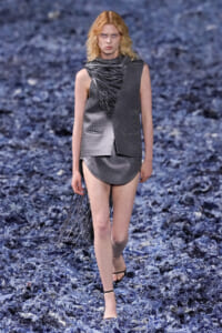 Model walking a runway in a gray plaid, sleeveless suit with an angular, fringed scarf draped over the shoulder and a short plaid skirt with a fringe train behind. Hard-soled sandals visible on a blue-textured runway surface.