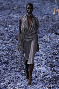 Model walking down a blue-toned runway in a gray fringe ensemble over a plaid midi skirt, dramatic and sculptural.