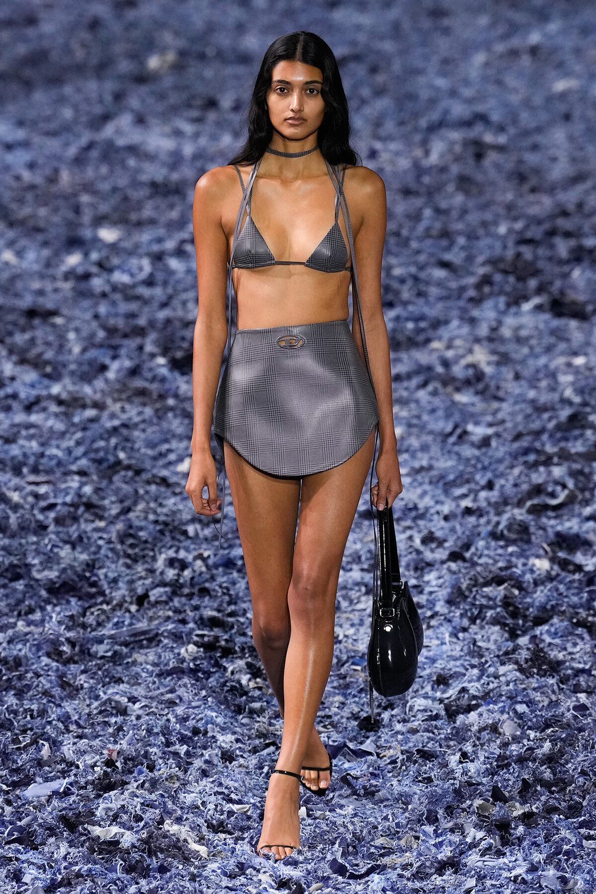 Model walks a runway in a gray plaid bikini top and matching high-waisted skirt, carrying a black handbag.
