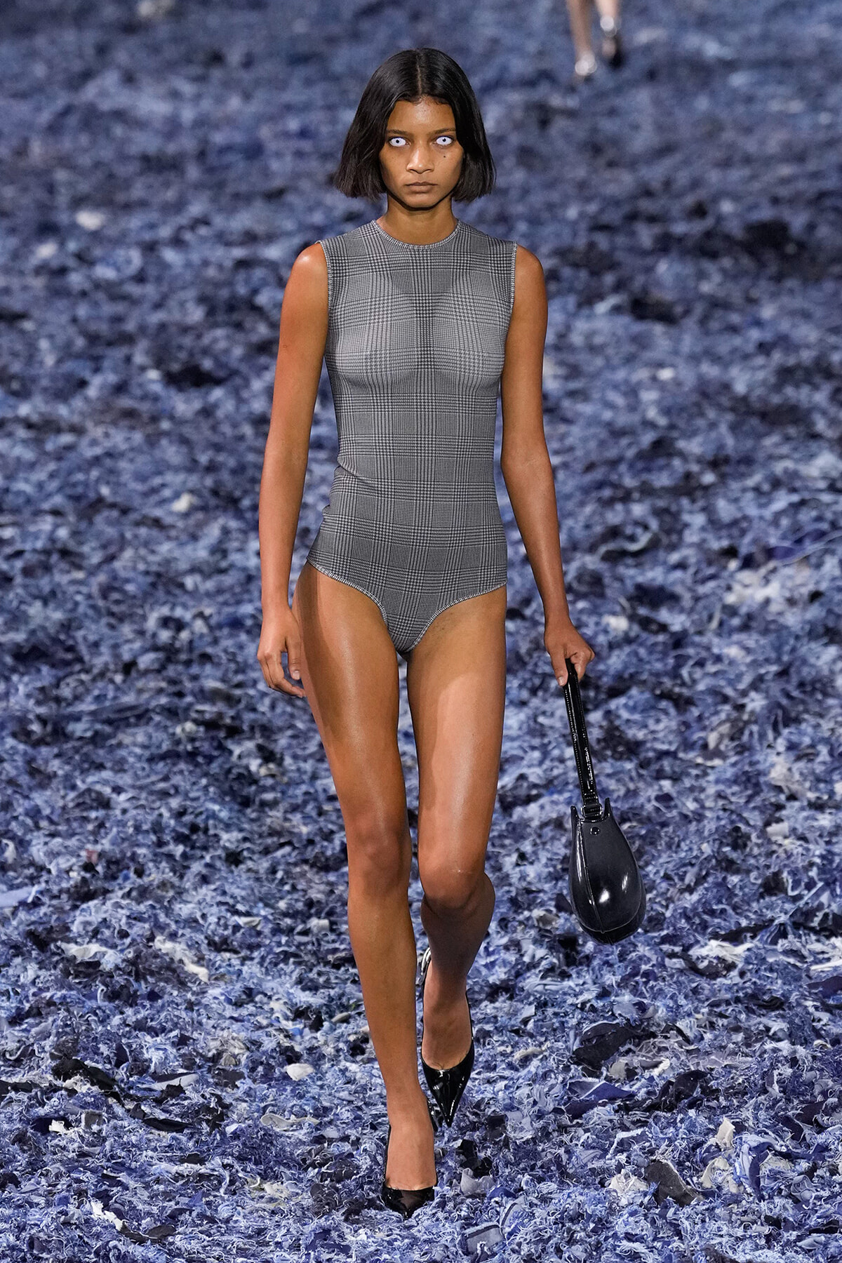 Model walks the runway in a gray plaid sleeveless bodysuit, black heels, and a small black handbag.