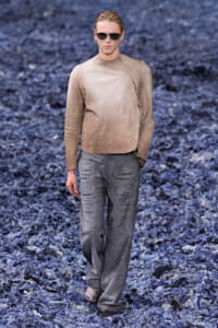 Male model walking a runway in a beige long-sleeve top and shimmery gray wide-leg pants, wearing sunglasses on a blue-toned floor