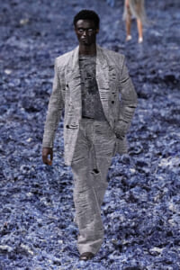 Male model walking a runway in a distressed light-gray suit with ripped detailing on a blue textured floor backdrop.