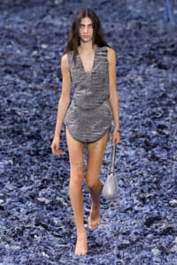 Model walking on a blue rocky runway wearing a gray plaid sleeveless romper with a matching belt and a small silver bag, barefoot on one footch? Note: avoid errors.