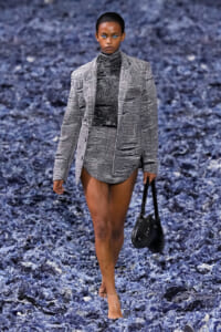 Model on a runway wearing a gray plaid blazer with matching shorts, carrying a black handbag on a blue textured backdrop.