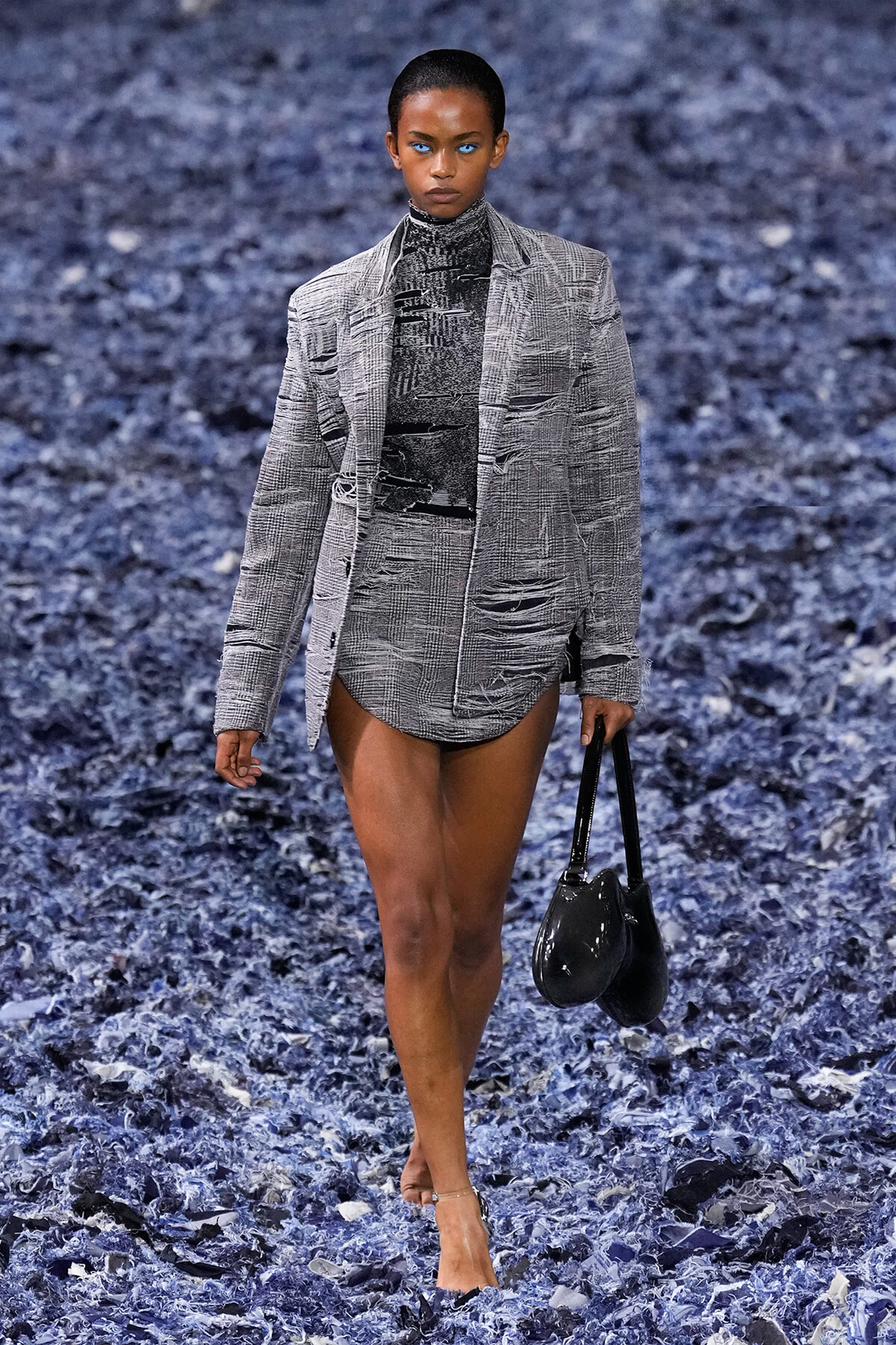 Model on a runway wearing a gray plaid blazer with matching shorts, carrying a black handbag on a blue textured backdrop.