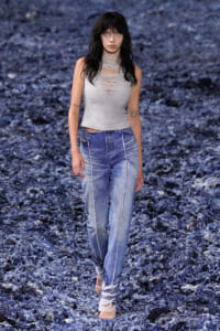 Model walking a runway in a light gray distressed sleeveless top and high-waisted blue jeans with white seams, barefoot with fabric wraps around ankles, against a textured blue backdrop.