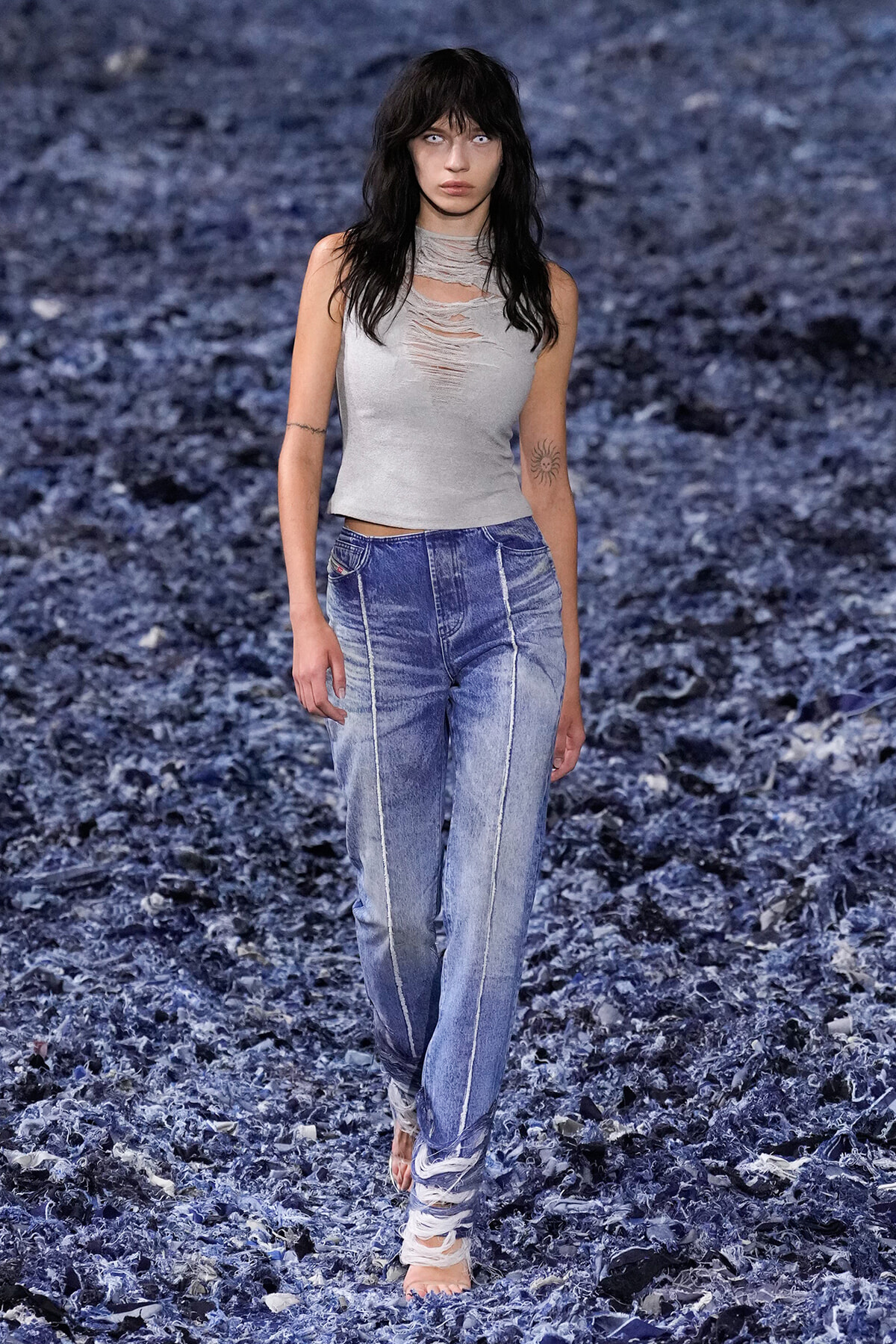 Model walking a runway in a light gray distressed sleeveless top and high-waisted blue jeans with white seams, barefoot with fabric wraps around ankles, against a textured blue backdrop.