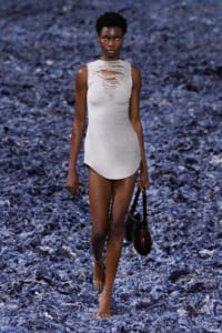 Model walking a blue-textured runway in a light gray distressed sleeveless dress, holding a dark handbag.