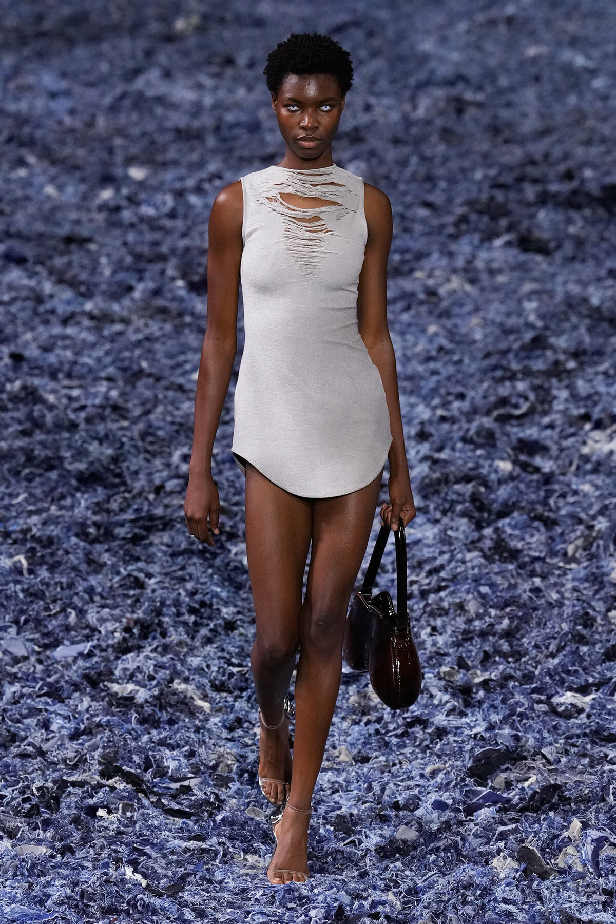 Model walking a blue-textured runway in a light gray distressed sleeveless dress, holding a dark handbag.