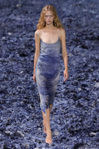 Model walking a runway in a light blue denim slip dress with a distressed, frayed hem, barefoot on a blue textured runway surface.