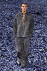 Male model wearing an oversized gray distressed sweatshirt and matching pants with large rips running down the chest and legs, walking a blue-textured runway background.