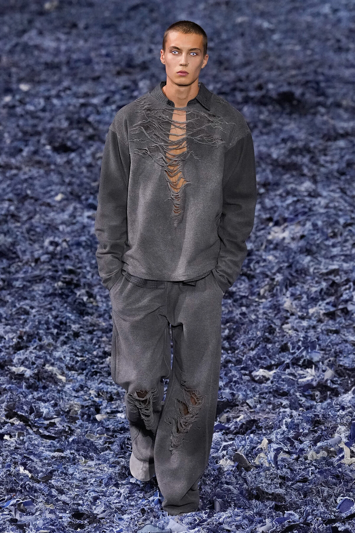 Male model wearing an oversized gray distressed sweatshirt and matching pants with large rips running down the chest and legs, walking a blue-textured runway background.