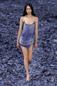 Model walking on a blue textured runway in a light-wash denim slip dress with spaghetti straps and fringe at the hem.
