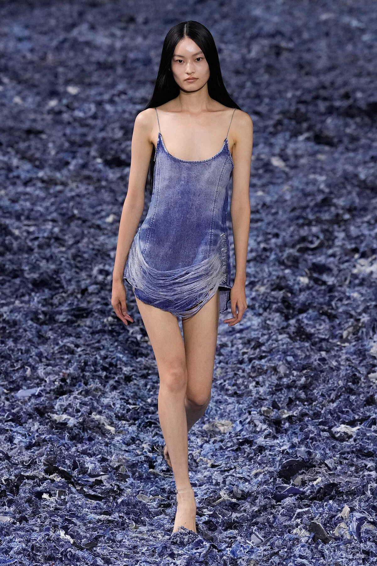 Model walking on a blue textured runway in a light-wash denim slip dress with spaghetti straps and fringe at the hem.