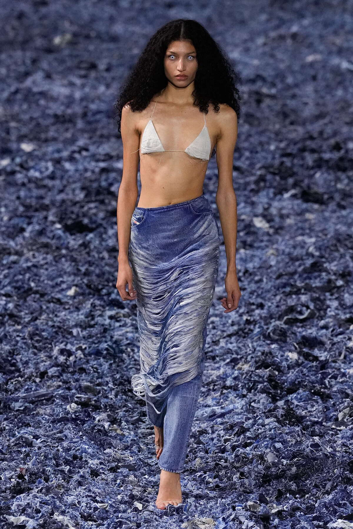 Model walking barefoot on a blue-toned rocky runway, wearing a beige triangle bikini top and a long, ruched blue denim skirt.